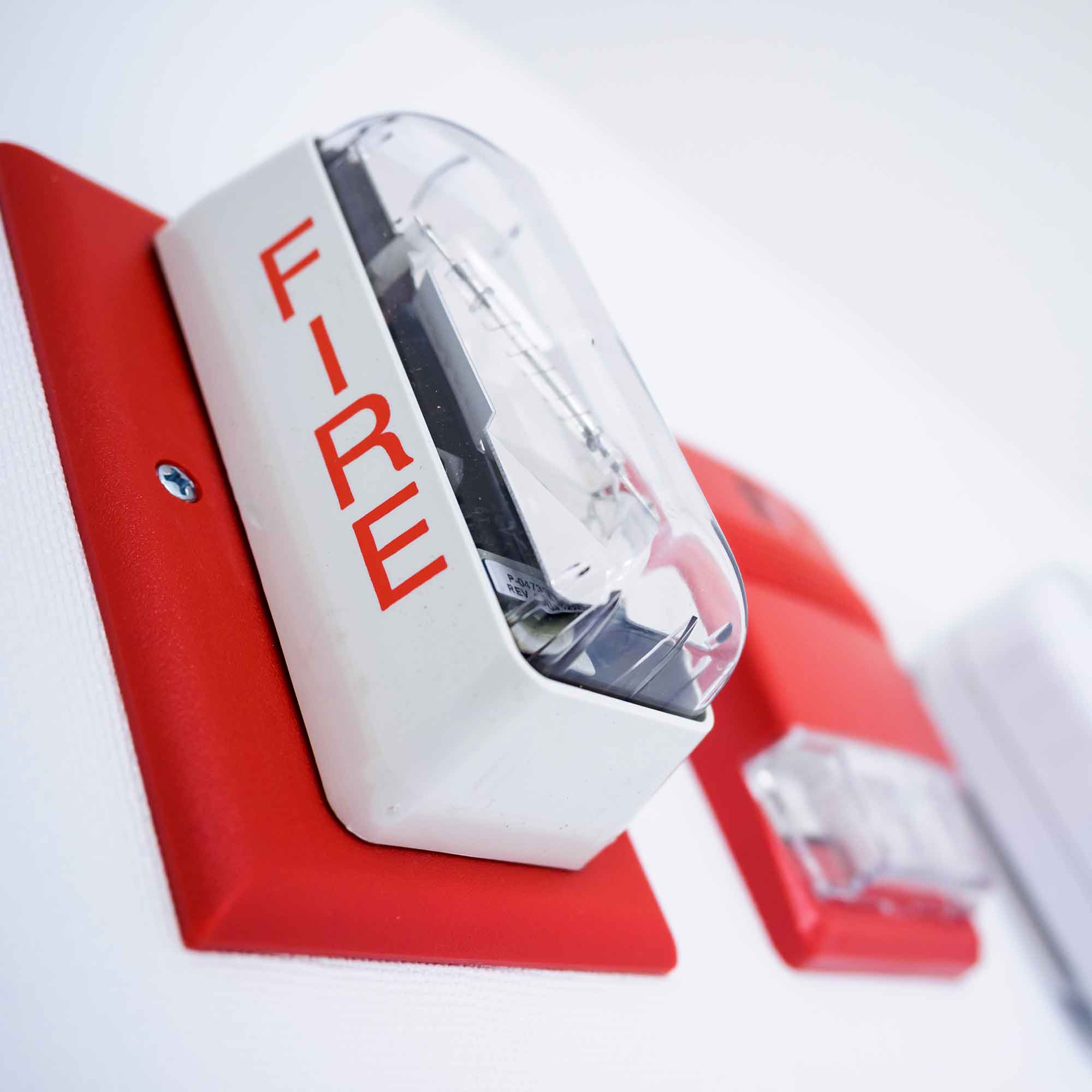 Close-up of a fire alarm with a red base and clear cover on a white wall.