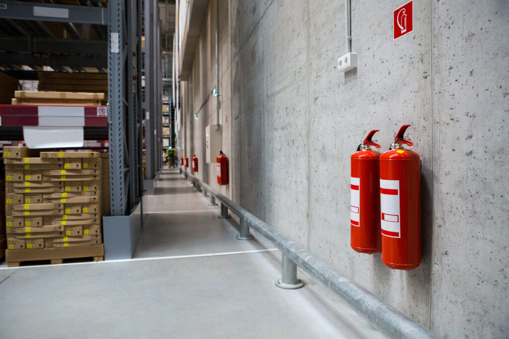 Fire extinguishers on a warehouse wall with stacked boxes and shelves in the background.
