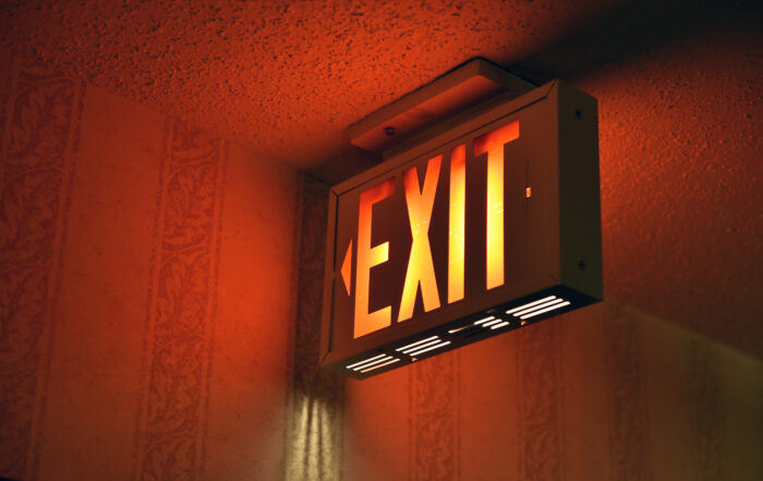 Illuminated exit sign with an arrow pointing left, mounted on a wall with a patterned wallpaper background.