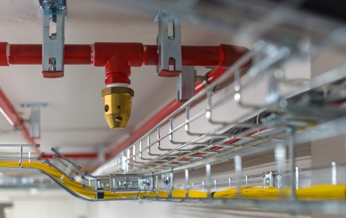 Close-up of a fire sprinkler head and red pipes above cable trays with yellow and gray electrical wires.