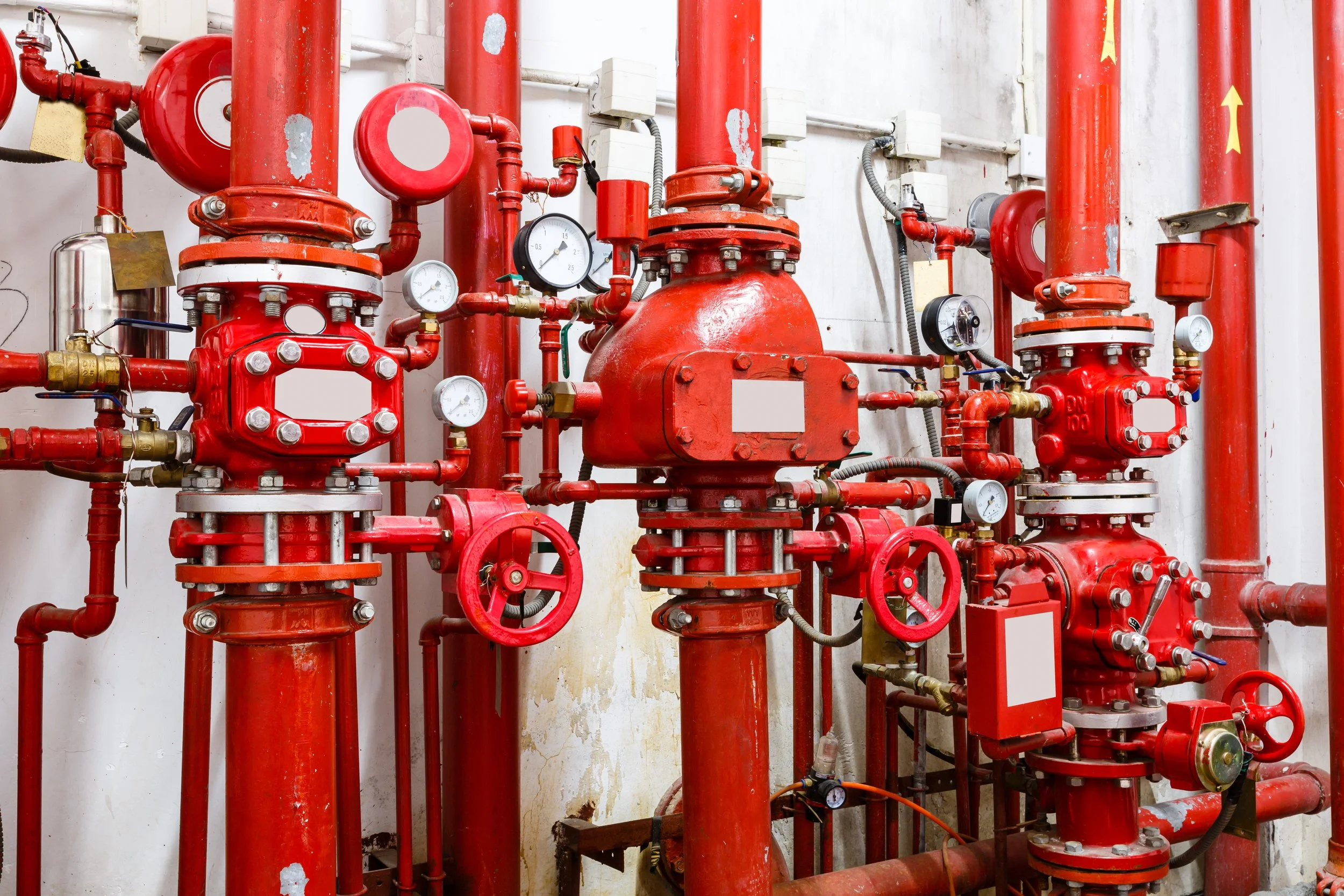Red industrial fire sprinkler system valves, pipes, and gauges mounted on a white wall in a mechanical room.