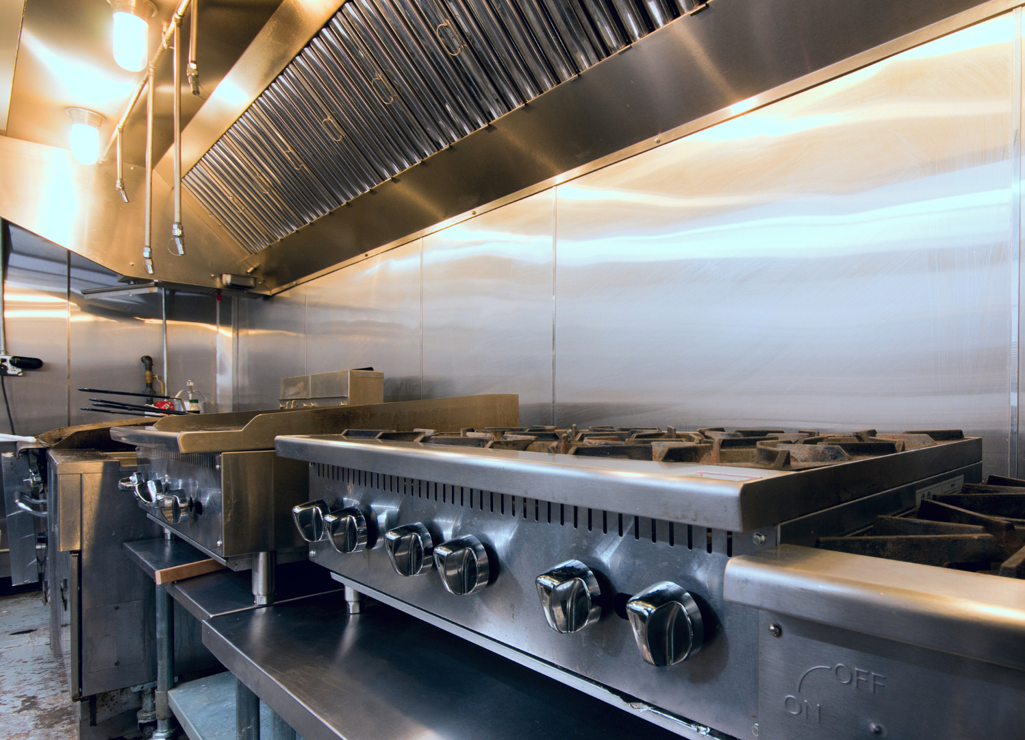 Commercial kitchen with stainless steel stoves, ovens, and ventilation hood under bright lighting.