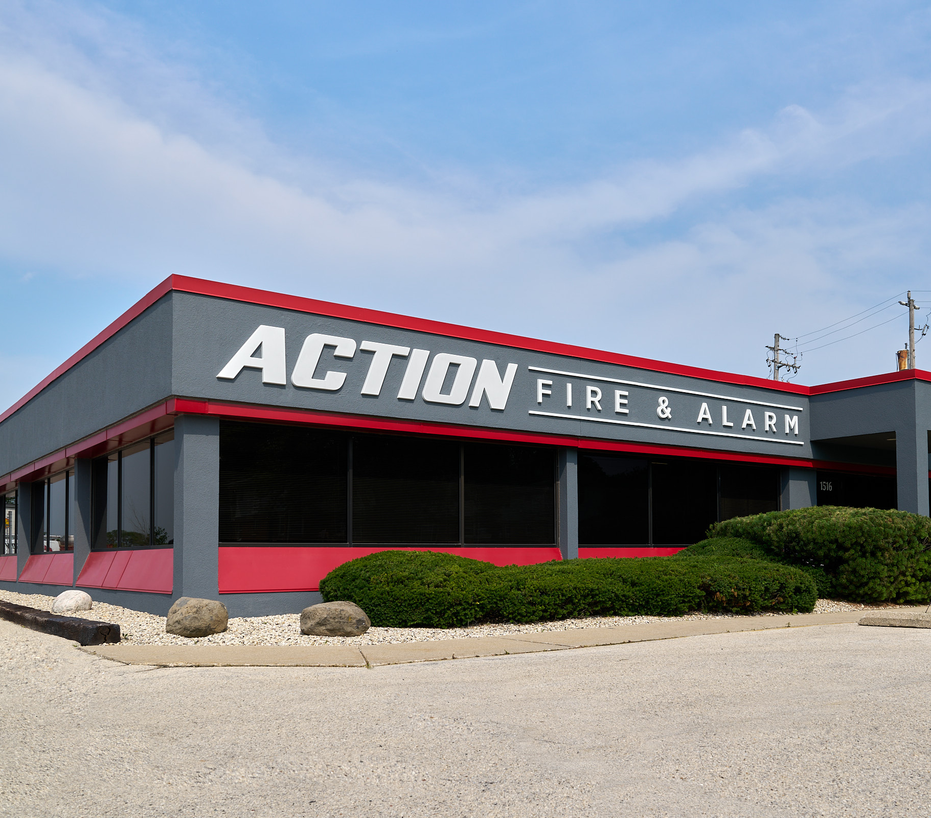 A gray and red building with a sign reading "Action Fire & Alarm" above large windows and shrubs.